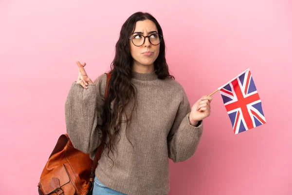 Young caucasian woman holding an United Kingdom flag isolated on pink background with fingers crossing and wishing the best