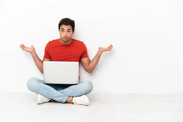 Caucasian handsome man with a laptop sitting on the floor having doubts while raising hands