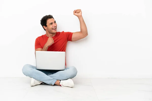 Caucasian handsome man with a laptop sitting on the floor celebrating a victory