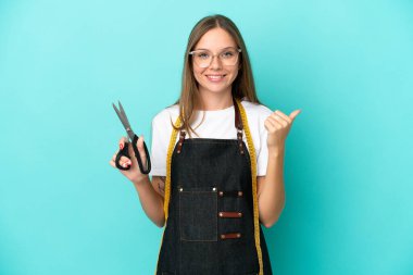 Genç Litvanyalı terzi kadın, mavi arka planda izole edilmiş bir şekilde bir ürün sunmak için yan tarafa bakıyor.