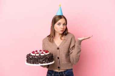 Litvanyalı genç bir kadın pembe arka planda izole edilmiş doğum günü pastasını tutarken ellerini kaldırırken şüphe duyuyor.