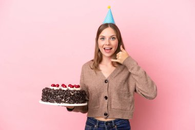 Elinde doğum günü pastası tutan Litvanyalı genç bir kadın pembe arka planda izole bir şekilde telefon jesti yapıyor. Bana geri dönüş işareti ver.