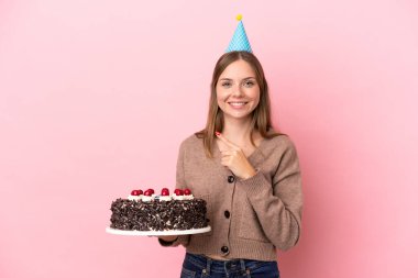 Elinde doğum günü pastası tutan Litvanyalı genç bir kadın pembe arka planda izole edilmiş bir şekilde bir ürünü sunmak için kenarı işaret ediyor.