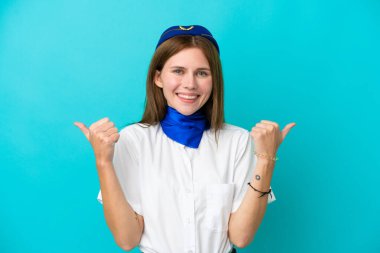 Uçak hostesi İngiliz kadın mavi arka planda izole edilmiş. Baş parmakları havada ve gülümsüyor.