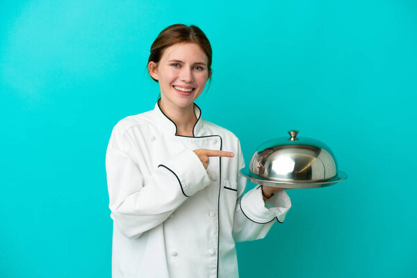 Young chef woman with tray isolated on blue background pointing to the side to present a product