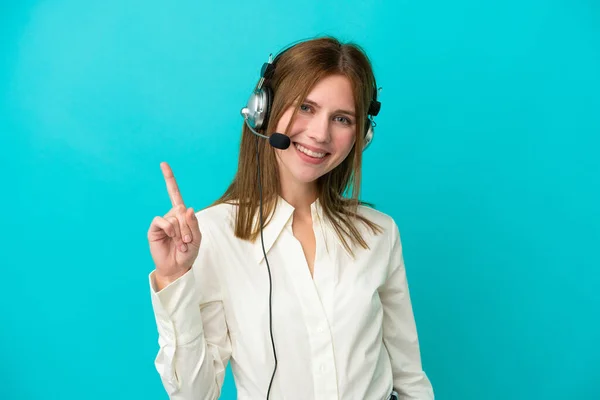 Telemarketer English woman working with a headset isolated on blue background showing and lifting a finger in sign of the best