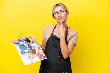 Kafkasyalı genç bir kadın sarı arka planda izole edilmiş bir palet tutuyor ve yukarı bakarken şüpheleri var.