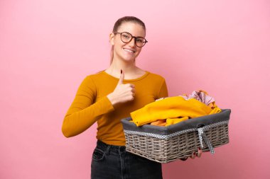 Pembe arka planda izole edilmiş çamaşır sepetini tutan beyaz bir kadın baş parmak işareti yapıyor.