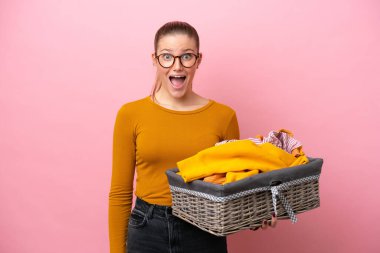 Pembe arka planda, yüzünde sürpriz bir ifadeyle izole edilmiş çamaşır sepeti tutan beyaz bir kadın.