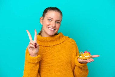 Mavi arka planda izole edilmiş turta tutan beyaz bir kadın gülümsüyor ve zafer işareti gösteriyor.