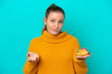 Mavi arka planda izole edilmiş turta tutan beyaz bir kadın omuzlarını kaldırırken şüphe uyandırıyor.