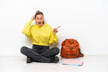 Beyaz arka planda beyaz bir zeminde oturan genç beyaz kadın şaşırdı ve parmağını yana çevirdi.