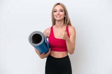 Genç sarışın kadın yoga derslerine giderken beyaz arka planda izole edilmiş bir minder tutuyor ve baş parmak hareketi yapıyor.