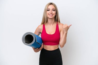 Genç sarışın kadın yoga derslerine giderken beyaz arka planda izole edilmiş bir minder tutuyor ve bir ürünü sunmak için yan tarafı işaret ediyor.