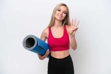 Genç sarışın kadın yoga derslerine giderken beyaz arka planda izole edilmiş bir minder tutuyor mutlu ve parmaklarıyla üçe kadar sayıyor.