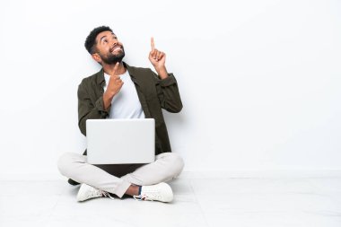 Yerde dizüstü bilgisayarı olan genç Brezilyalı bir adam işaret parmağıyla beyaz üzerine izole edilmiş olarak oturuyor. Harika bir fikir.