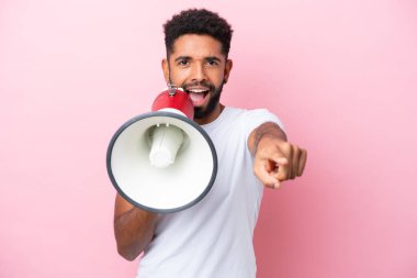 Young Brazilian man isolated on pink background shouting through a megaphone to announce something while pointing to the front