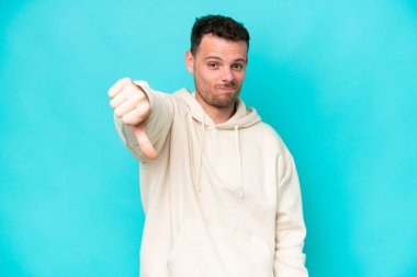 Mavi arka planda soyutlanmış beyaz, yakışıklı genç bir adam, olumsuz bir ifadeyle baş parmağını aşağı gösteriyor.