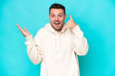 Beyaz tenli yakışıklı bir adam mavi arka planda izole edilmiş telefon jestleri yapıyor.