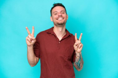 Mavi arka planda izole edilmiş beyaz, yakışıklı genç bir adam iki eliyle zafer işareti gösteriyor.