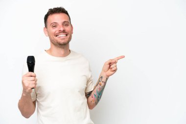 Young caucasian singer man picking up a microphone isolated on white background pointing finger to the side