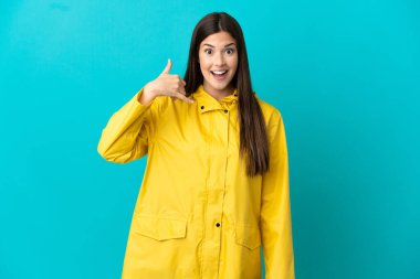 Teenager Brazilian girl wearing a rainproof coat over isolated blue background making phone gesture. Call me back sign