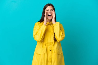Teenager Brazilian girl wearing a rainproof coat over isolated blue background shouting and announcing something