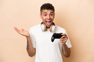 Young Brazilian man playing with a video game controller isolated on beige background with shocked facial expression