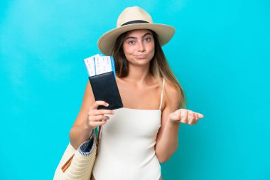 Young woman in swimsuit holding passport isolated on blue background making doubts gesture while lifting the shoulders