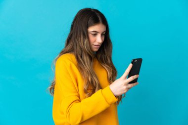 Mavi arka planda izole edilmiş genç beyaz kadın cep telefonuyla mesaj ya da e-posta gönderiyor.