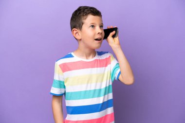 Little boy isolated on purple background keeping a conversation with the mobile phone