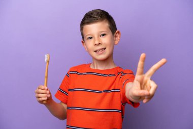 Little boy brushing teeth isolated on purple background smiling and showing victory sign