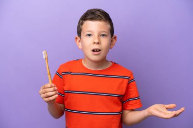 Little boy brushing teeth isolated on purple background with shocked facial expression