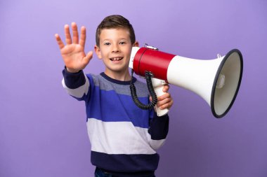 Little boy isolated on purple background holding a megaphone and saluting with hand with happy expression