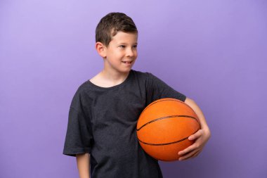 Mor arka planda izole edilmiş basketbol oynayan küçük çocuk yana bakıyor ve gülümsüyor.