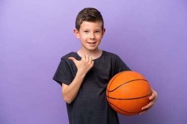 Little boy playing basketball isolated on purple background pointing to the side to present a product