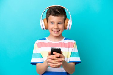 Little boy isolated on blue background listening music with a mobile and looking front