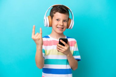 Little boy isolated on blue background listening music with a mobile making rock gesture