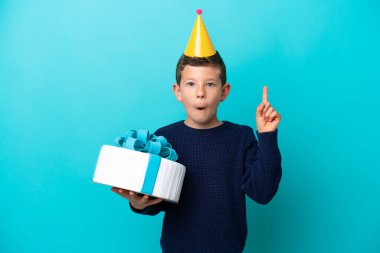Küçük çocuk mavi arka planda izole edilmiş doğum günü pastasını tutarken bir parmağını kaldırarak çözümü bulmayı umuyor.