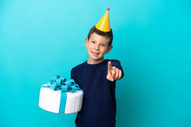 Little boy holding birthday cake isolated on blue background showing and lifting a finger