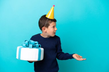 Little boy holding birthday cake isolated on blue background with surprise expression while looking side