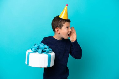 Little boy holding birthday cake isolated on blue background shouting with mouth wide open to the side
