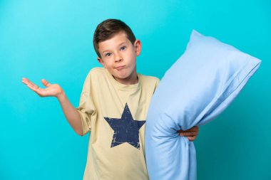 Little boy in pajamas isolated on blue background having doubts while raising hands