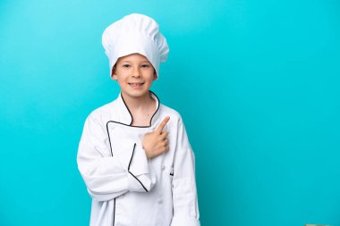 Mavi arka planda izole edilmiş küçük aşçı çocuk bir ürün sunmak için kenarı işaret ediyor.