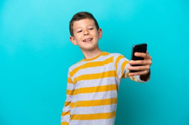 Mavi arka planda izole edilmiş küçük bir çocuk cep telefonuyla selfie çekiyor.