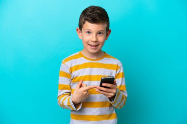 Little boy isolated on blue background using mobile phone and pointing it