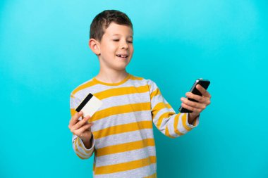 Küçük çocuk mavi arka planda izole edilmiş, kredi kartıyla cep telefonuyla alışveriş yapıyor.