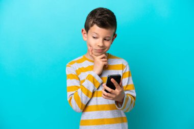 Little boy isolated on blue background using mobile phone and thinking