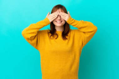 Kızıl saçlı kız mavi arka planda izole edilmiş gözleri elleriyle kaplayan ve gülümseyen.