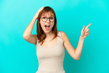 Redhead girl isolated on blue background surprised and pointing finger to the side